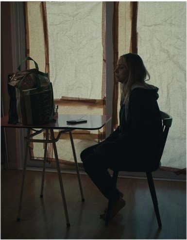 Maddy sitting alone at a table beside a curtained window
